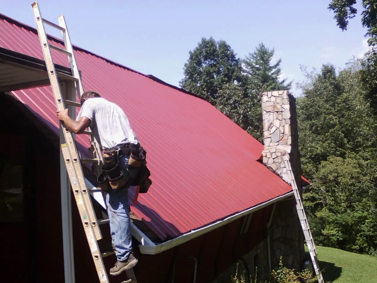 Roofer on a ladder beside a red metal roof and stone chimney — Soffit & Fascia Repair by Roofing Brothers in Macomb